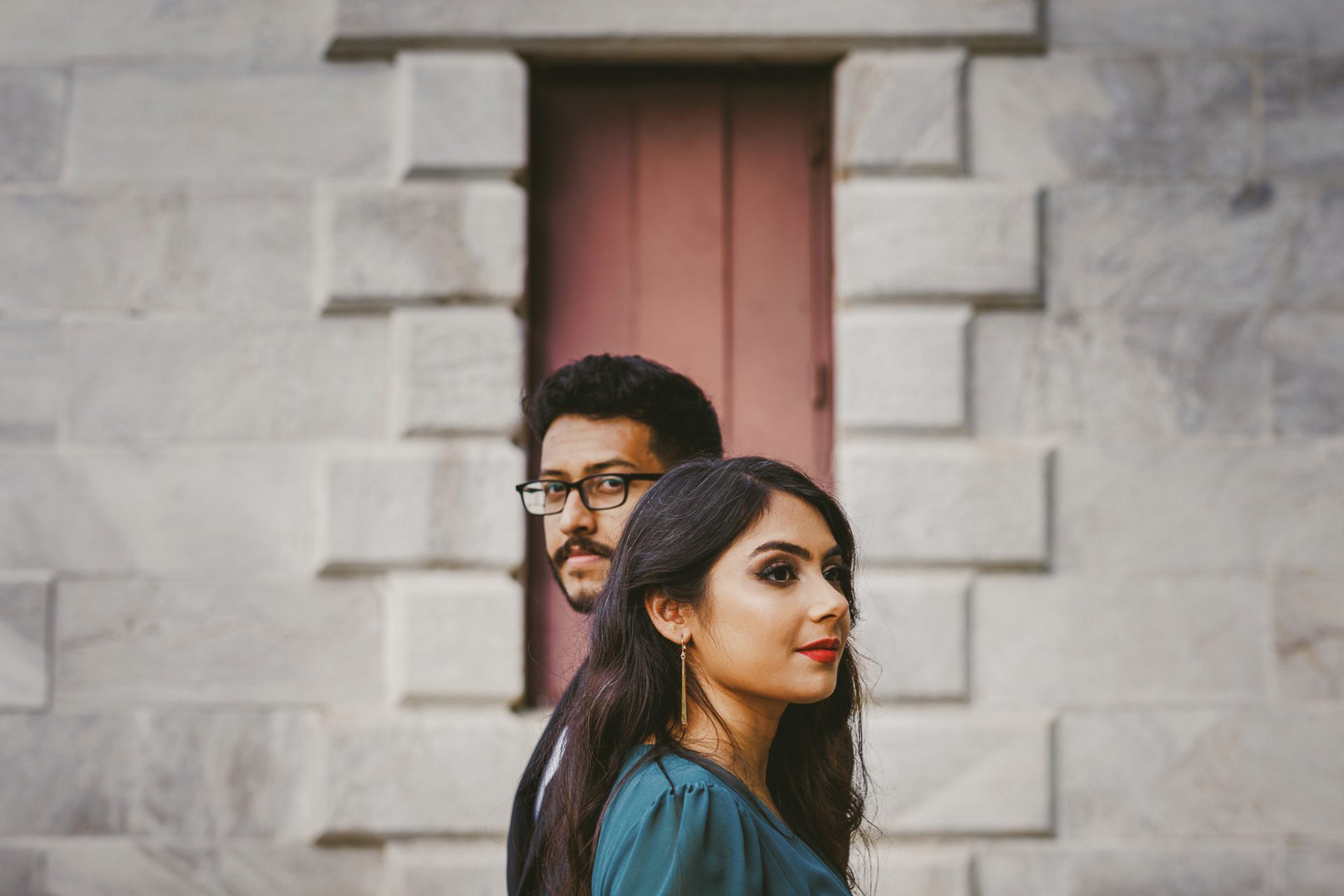 A man and woman stand sideways in front of a brick wall with a window. The man wears glasses and a black jacket, while the woman has long dark hair and wears a teal top. Both look towards the camera, capturing a candid moment perfect for any wedding photography portfolio.