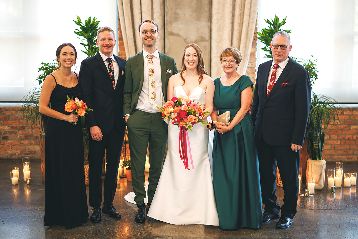 A wedding group photo featuring a bride and groom with four family members, smiling in a candlelit room with brick walls and green plants.