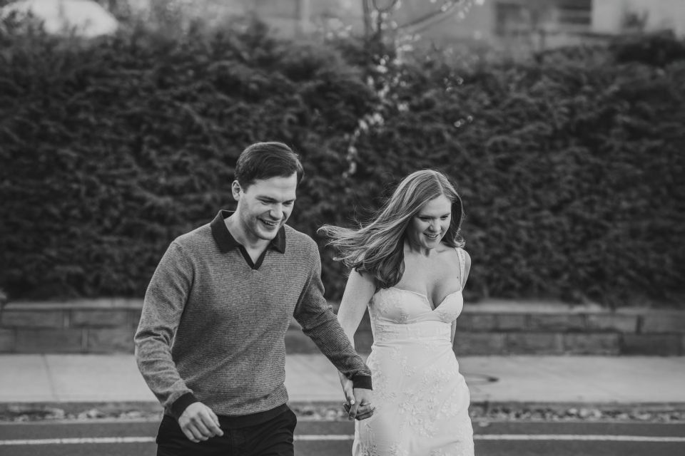 A couple holding hands and walking outdoors, both smiling. The woman is wearing a strapless dress, and the man is in a sweater. The background features shrubs and a sidewalk.