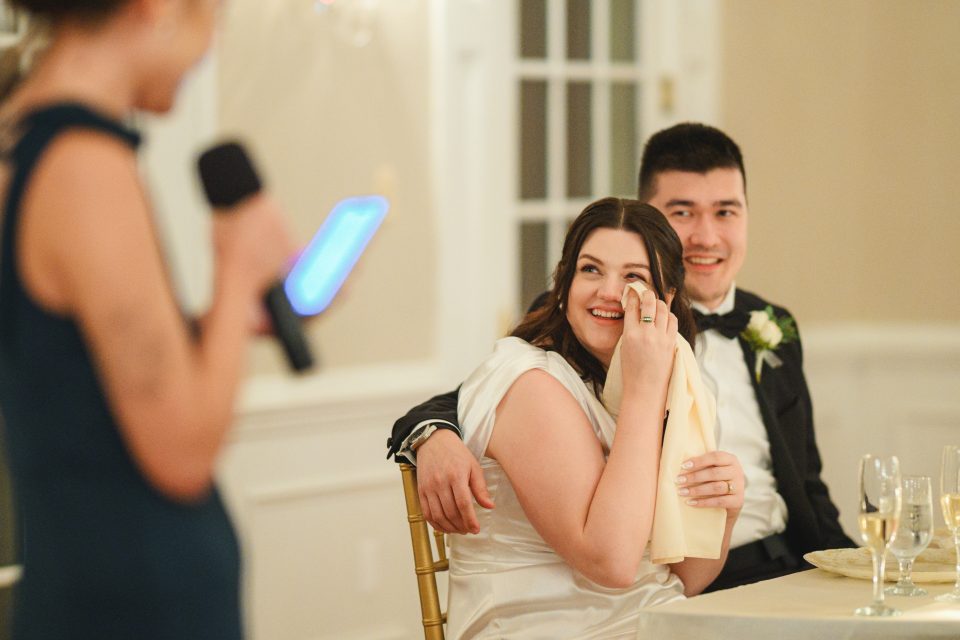 A couple sits at a table during a speech, with the woman holding a napkin to her face, smiling.