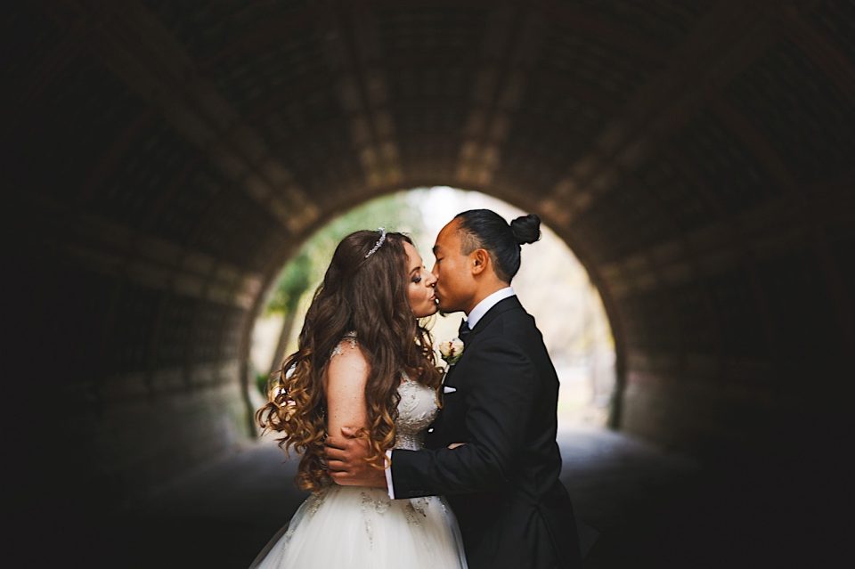 Ash + John, a bride and groom, kissing tenderly in a dimly lit tunnel, highlighted by natural light at the tunnel entrance.