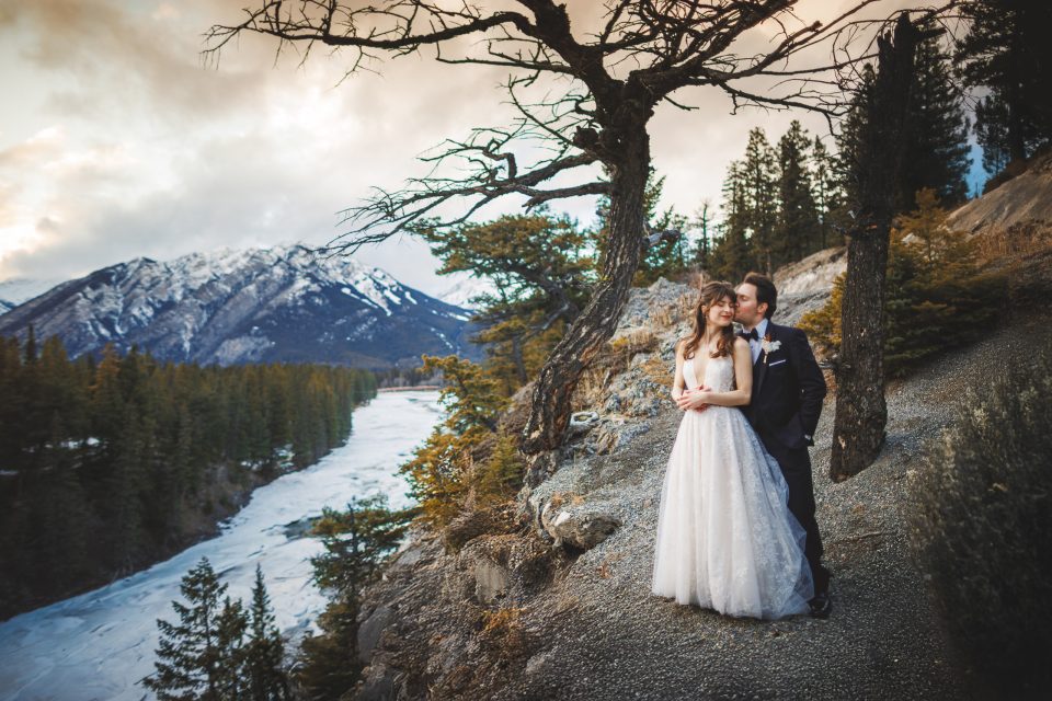 A bride and groom stand on a rocky ledge overlooking a snowy river and pine trees, with mountains in the background under a cloudy sky.