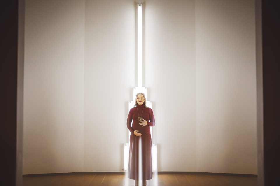 A person in a long red dress stands in front of a vertical light installation on a white wall, with hands resting on their midsection.