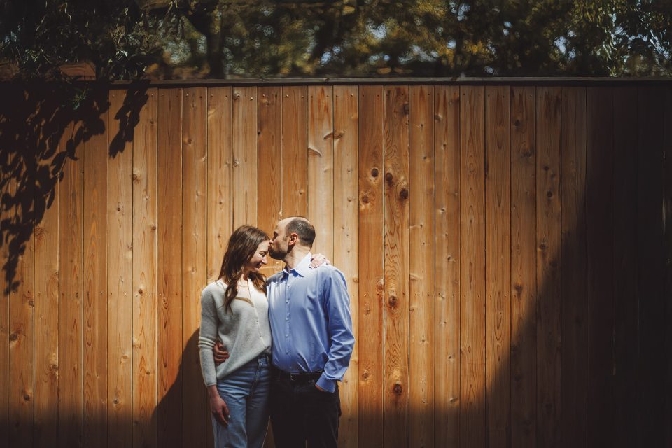 A couple stands close together in front of a wooden fence, with the man kissing the woman's forehead. Sunlight casts a shadow on part of the scene.