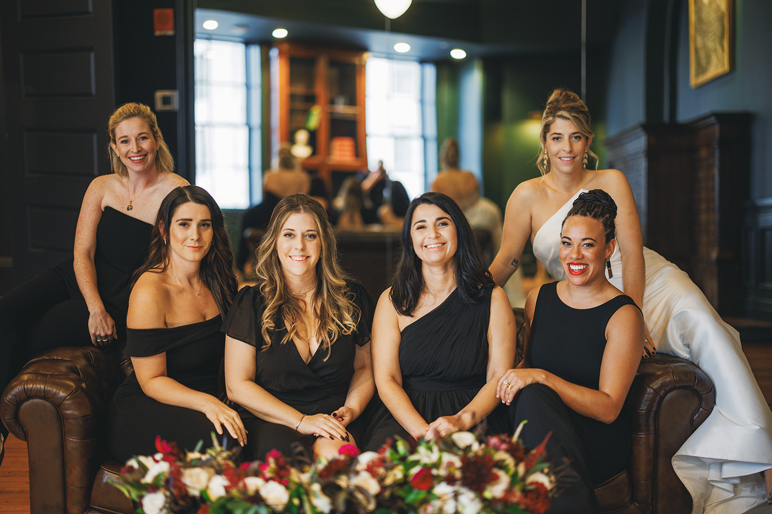 Six women, dressed in formal attire, pose together in a room with dark green walls.