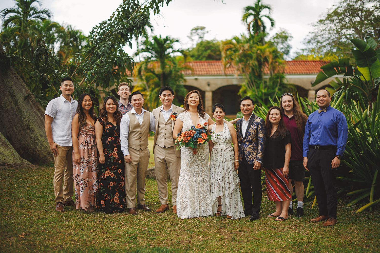 A group photo of people posing outdoors, with a couple in wedding attire at the center. The individuals are standing on grass, with trees and a building in the background.